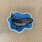 A die-cut sticker featuring an illustrated trout on a blue bubbly background, placed on a brown surface with a black polka-dot pattern.