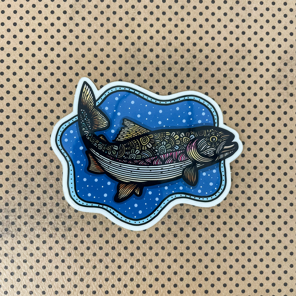 A die-cut sticker featuring an illustrated trout on a blue bubbly background, placed on a brown surface with a black polka-dot pattern.