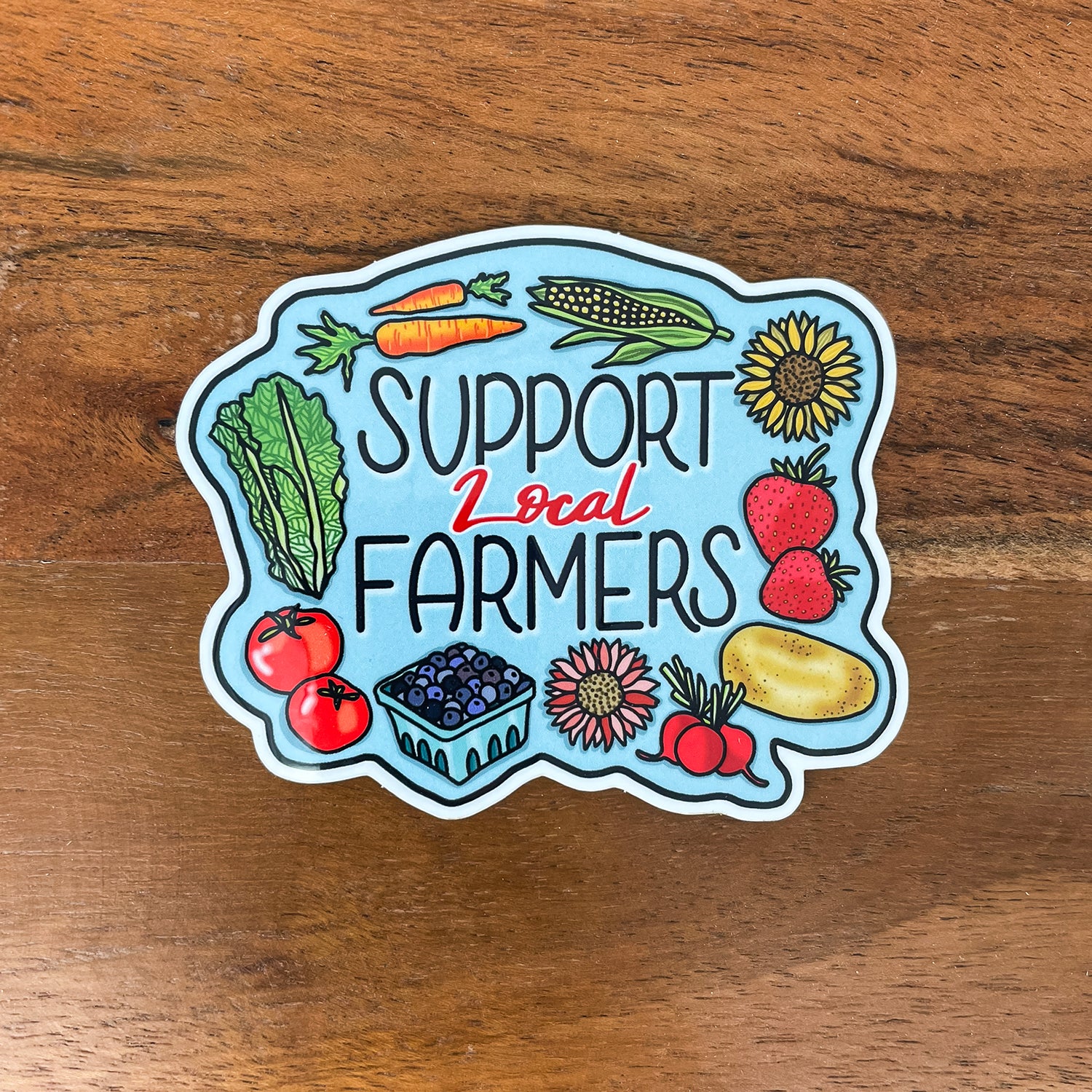 A die-cut sticker featuring the text 'SUPPORT Local FARMERS' surrounded by colorful illustrations of fresh vegetables and fruits, placed on a wooden surface.