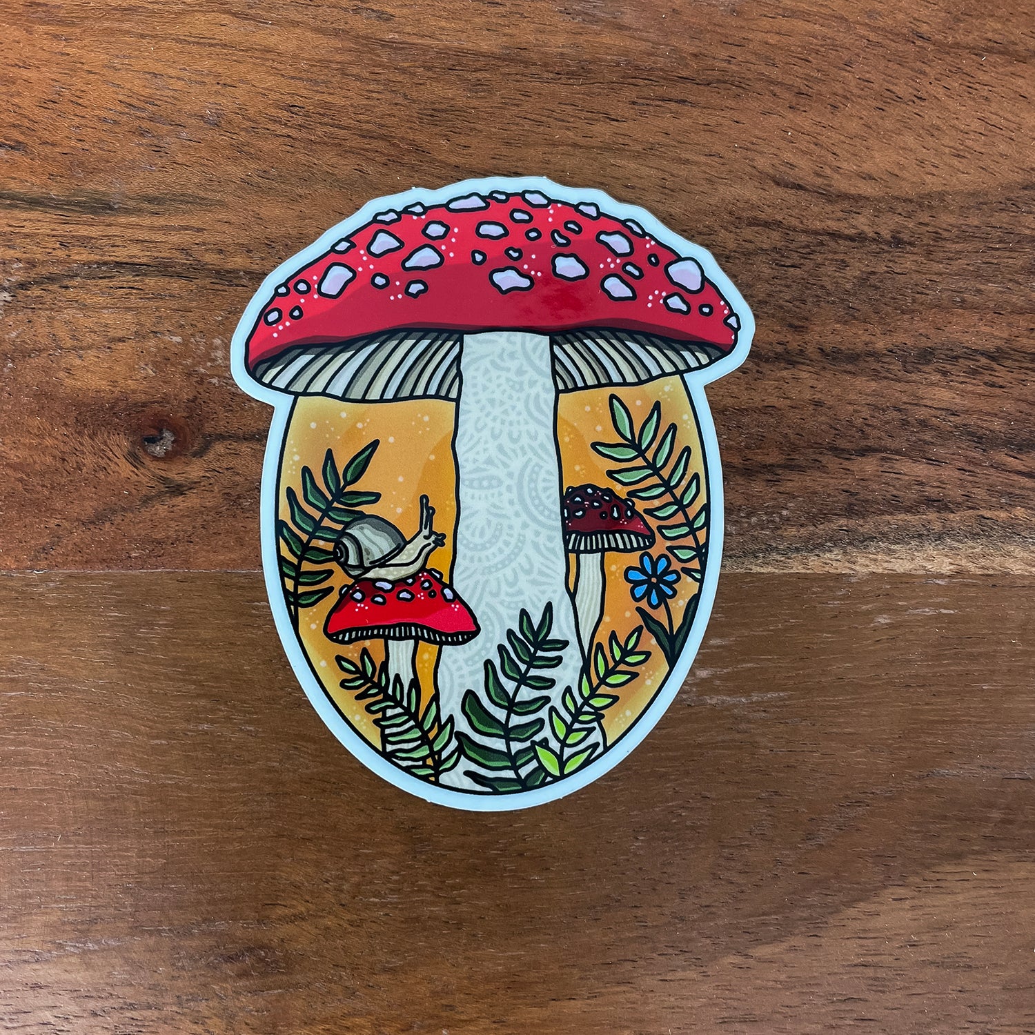 A die-cut sticker featuring a whimsical mushroom forest illustration, placed on a brown wooden surface. The sticker shows a large red-and-white mushroom, smaller mushrooms, green ferns, a snail, and a small blue flower, outlined in white.