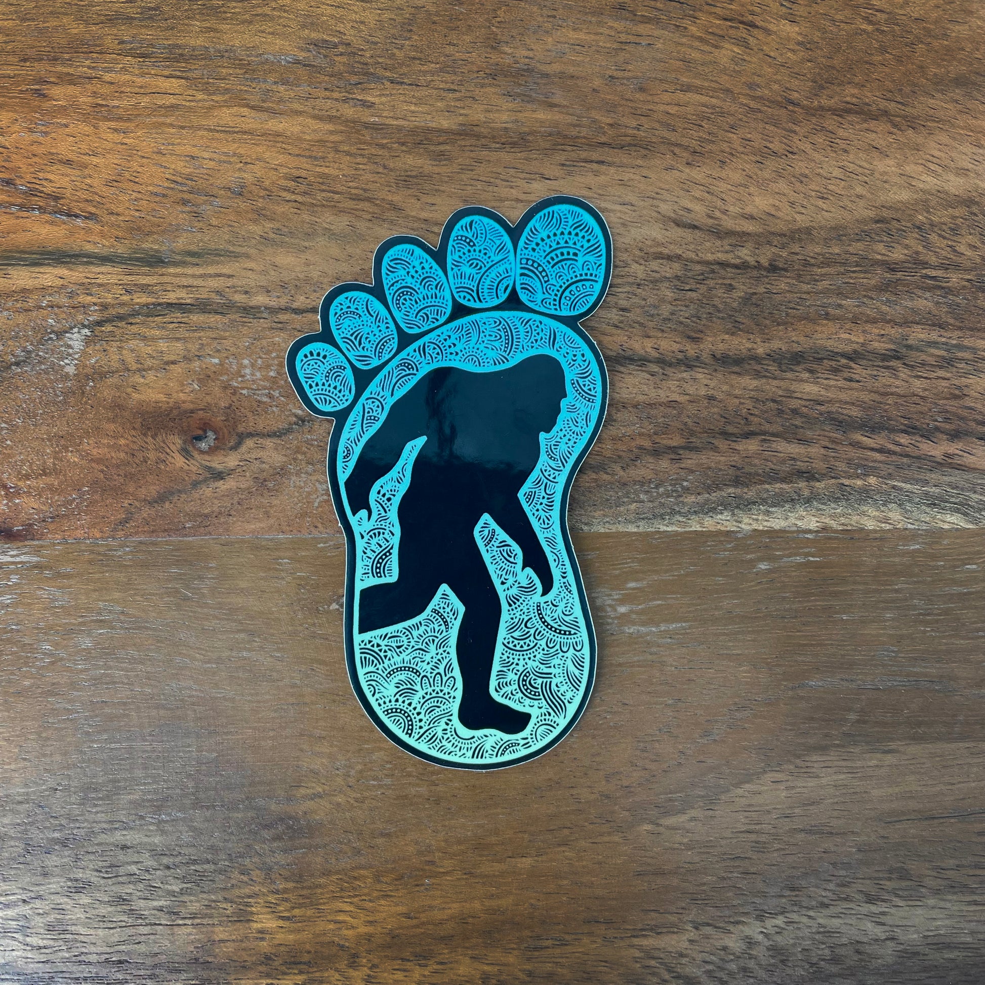 Sticker of a Bigfoot silhouette walking inside a foot shape laying on a wooden surface.