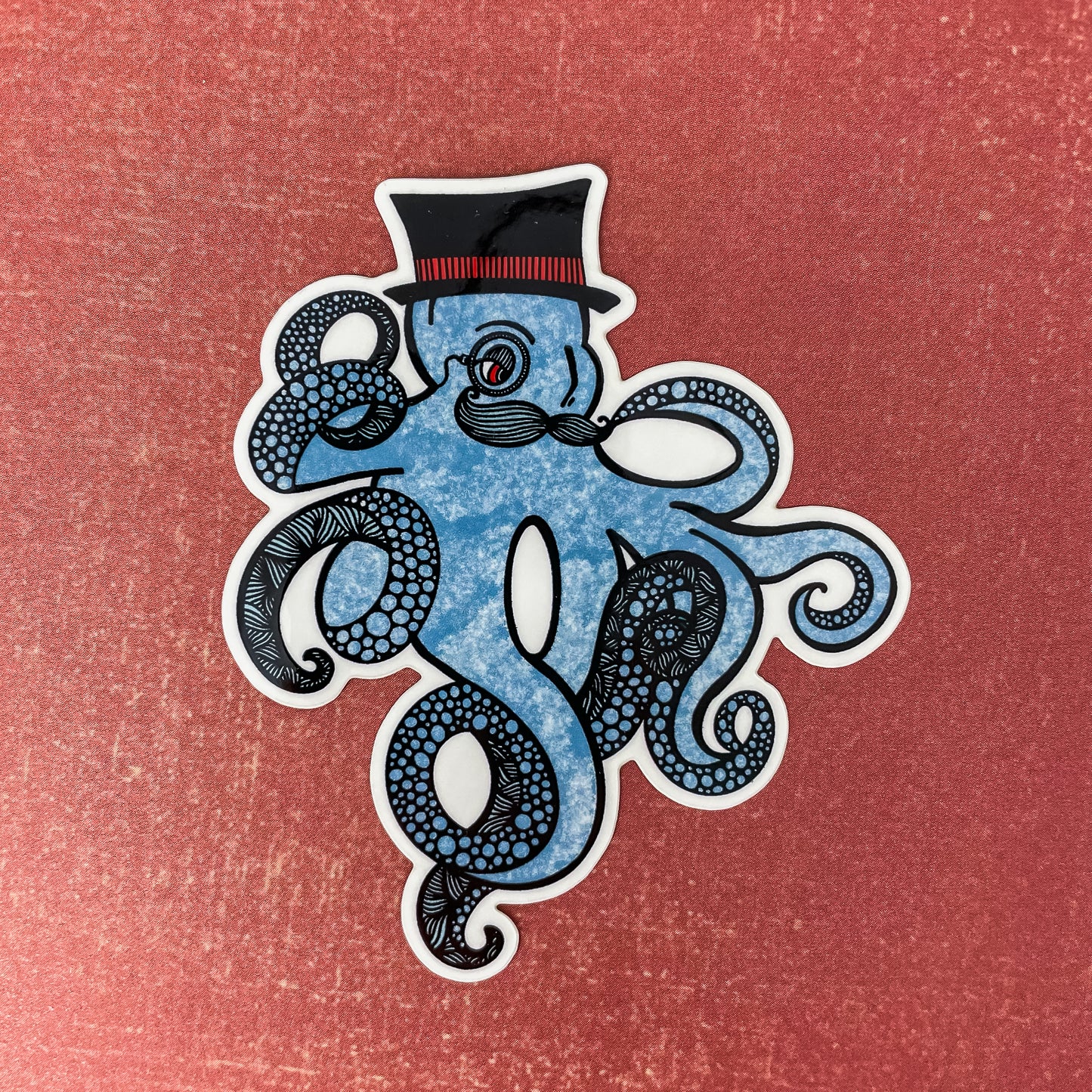 Blue kraken/octopus with a top hat and mustache. Detailed illustration made into a vinyl sticker laying on a textured red surface.