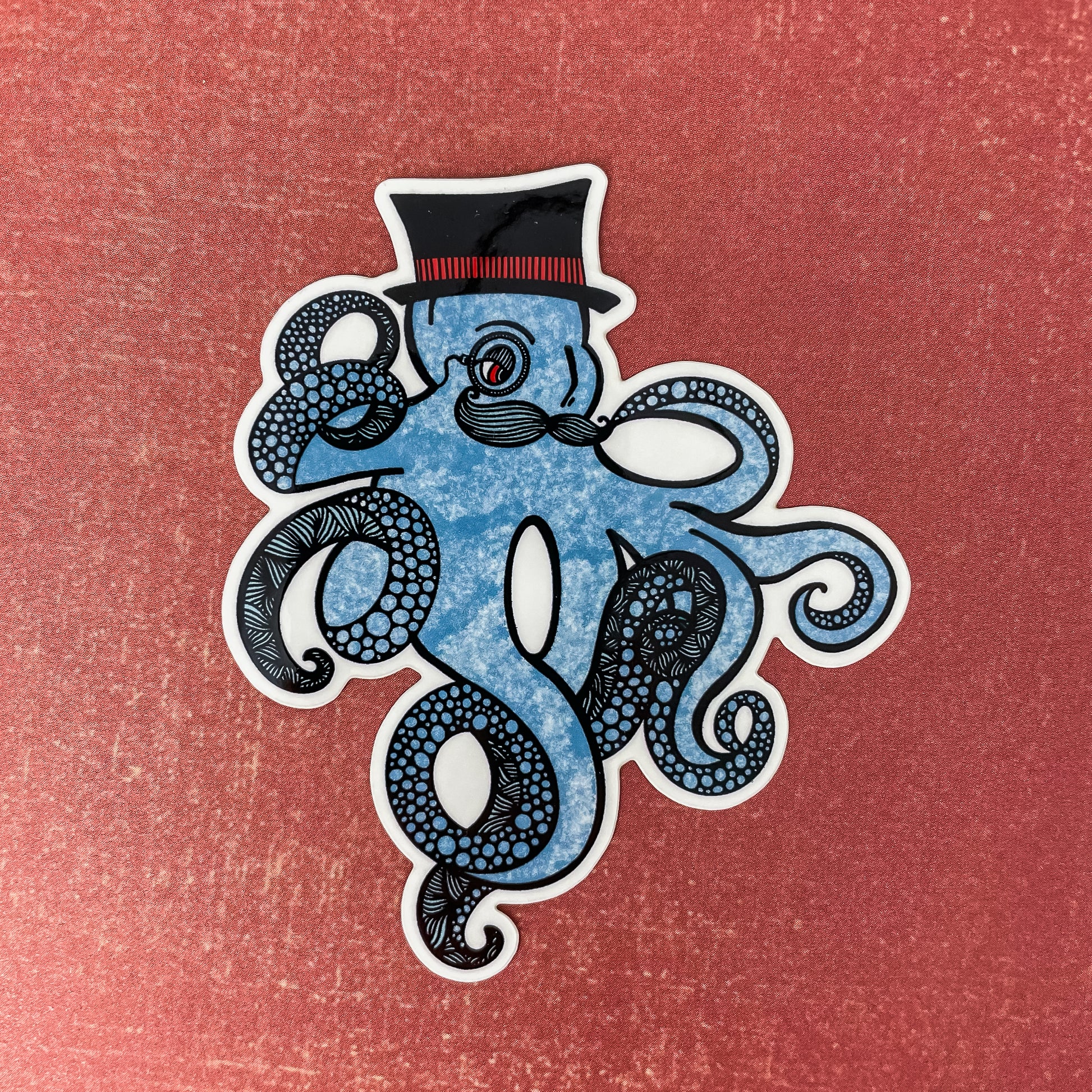 Blue kraken/octopus with a top hat and mustache. Detailed illustration made into a vinyl sticker laying on a textured red surface.