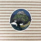 A circular sticker featuring an orca whale in front of a mountain under a starry night sky, with the word 'WASHINGTON' on a banner, shown on a beige and brown striped fabric background.