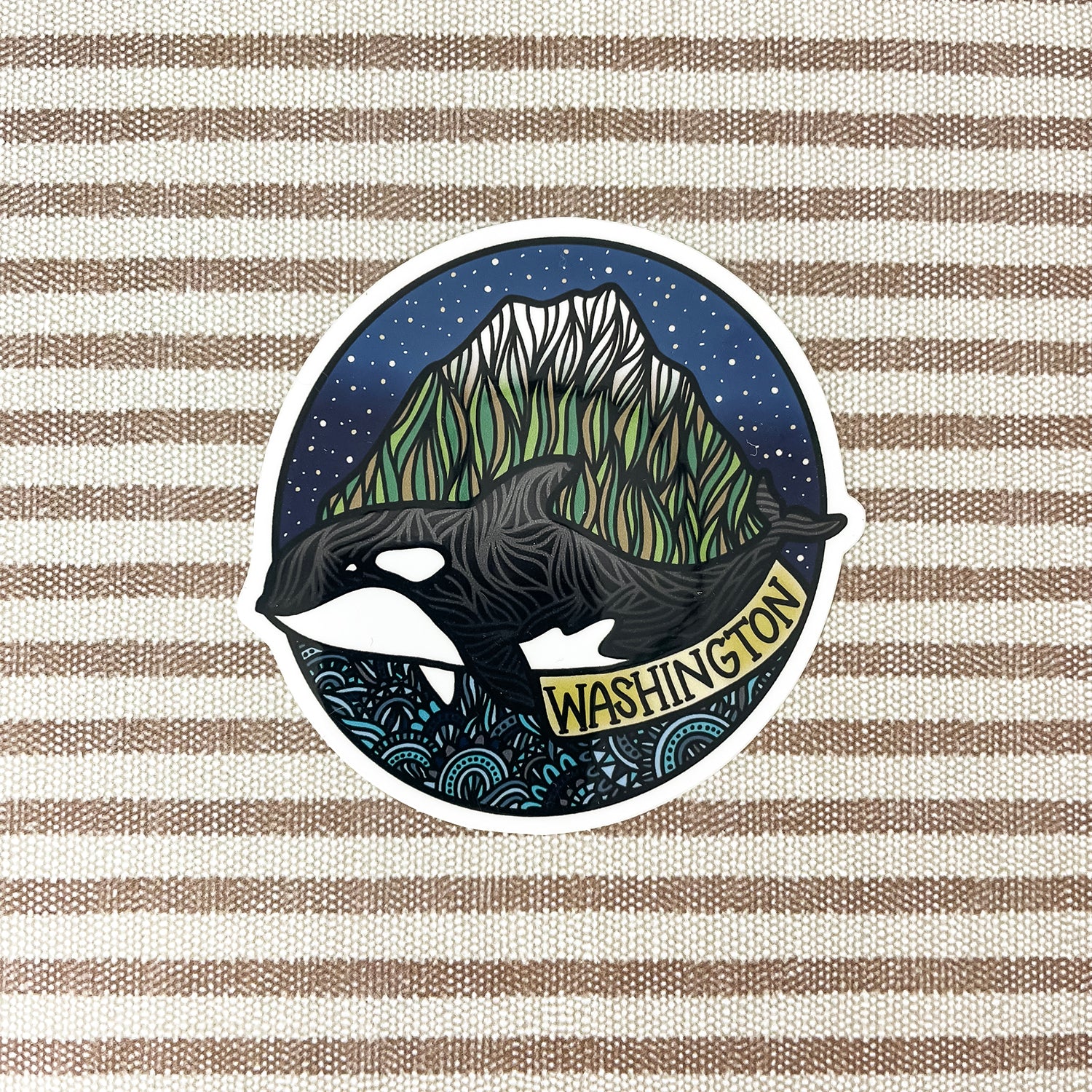 A circular sticker featuring an orca whale in front of a mountain under a starry night sky, with the word 'WASHINGTON' on a banner, shown on a beige and brown striped fabric background.