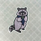 A die-cut sticker featuring a stylized raccoon wearing a blue striped scarf and holding a mug with the letters 'PNW' on it. The raccoon is decorated with intricate black floral patterns on a light gray base, set against a textured light gray background with a subtle diamond pattern.