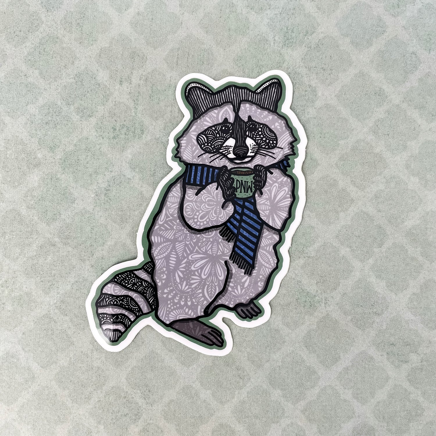 A die-cut sticker featuring a stylized raccoon wearing a blue striped scarf and holding a mug with the letters 'PNW' on it. The raccoon is decorated with intricate black floral patterns on a light gray base, set against a textured light gray background with a subtle diamond pattern.