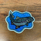 A die-cut sticker featuring an illustrated fish over a blue patterned background, placed on a wooden surface.