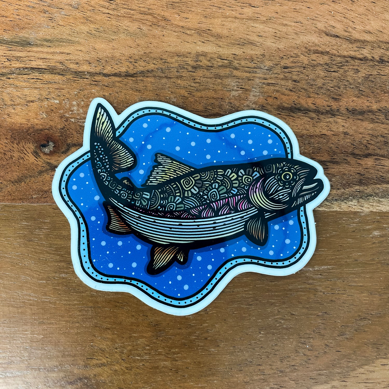 A die-cut sticker featuring an illustrated fish over a blue patterned background, placed on a wooden surface.