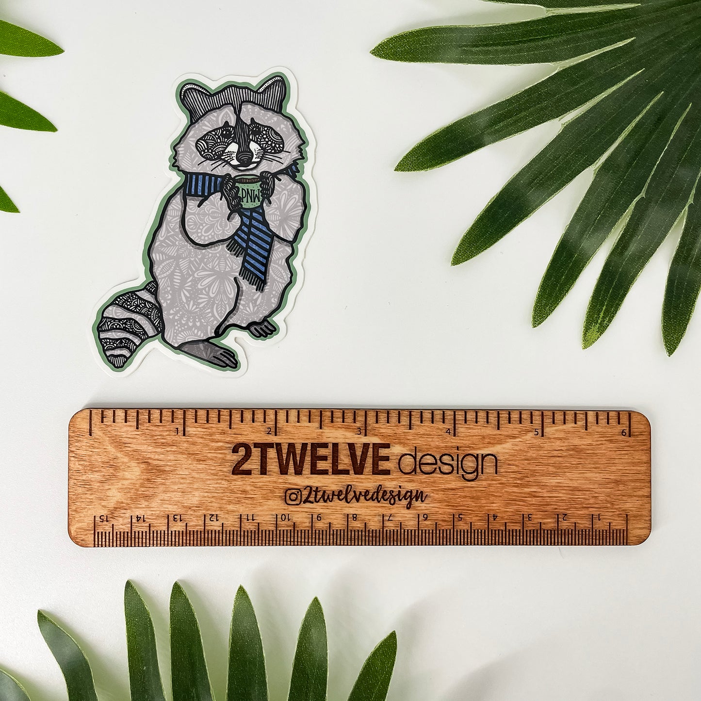 A die-cut sticker of a raccoon wearing a blue striped sweater and holding a PNW coffee mug, displayed next to a wooden ruler with "2Twelve Design" to show size, laying on a white background with green palm leaves.