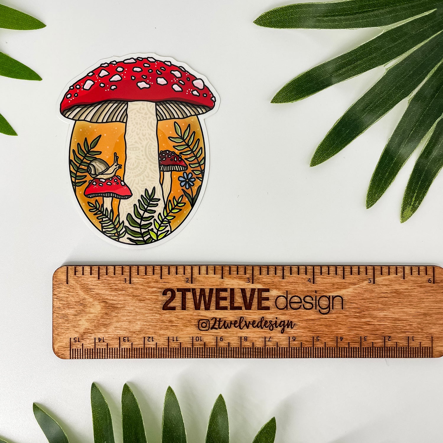 A die-cut sticker featuring a whimsical mushroom forest illustration with a small snail. Placed above a wooden ruler labeled 2Twelve Design. The scene is styled with green palm leaves on a white background.