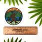 Circular sticker with a stylized mountain and bigfoot illustration. Laying on a white background with leaves surrounding and a ruler to show actual size.