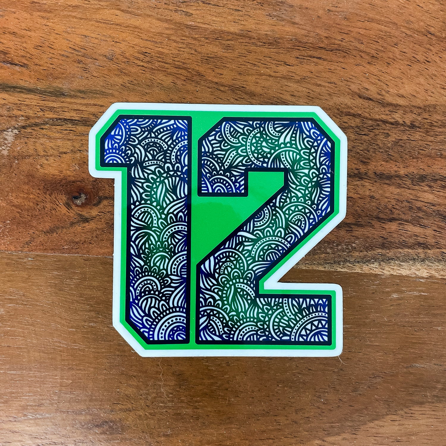 A die-cut sticker featuring the number 12 filled with a green, blue, and white mandala pattern, placed on a wooden surface.