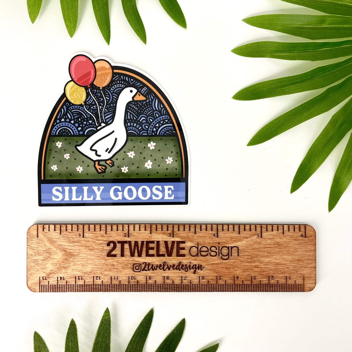 Sticker of a silly goose with balloons laying on a white background with leaves and a ruler to show actual size.