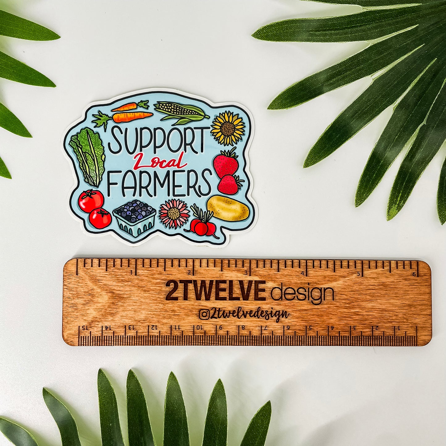 A die-cut sticker featuring an illustrated farm scene with vegetables, fruits, and the text 'SUPPORT Local FARMERS' is displayed above a wooden ruler. The sticker is photographed on a white surface with green palm leaves around it.