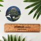 A circular sticker featuring an orca whale in front of a mountain range labeled 'WASHINGTON' is displayed on a white surface next to a wooden ruler and green palm leaves. The sticker is branded with '2Twelve Design'.