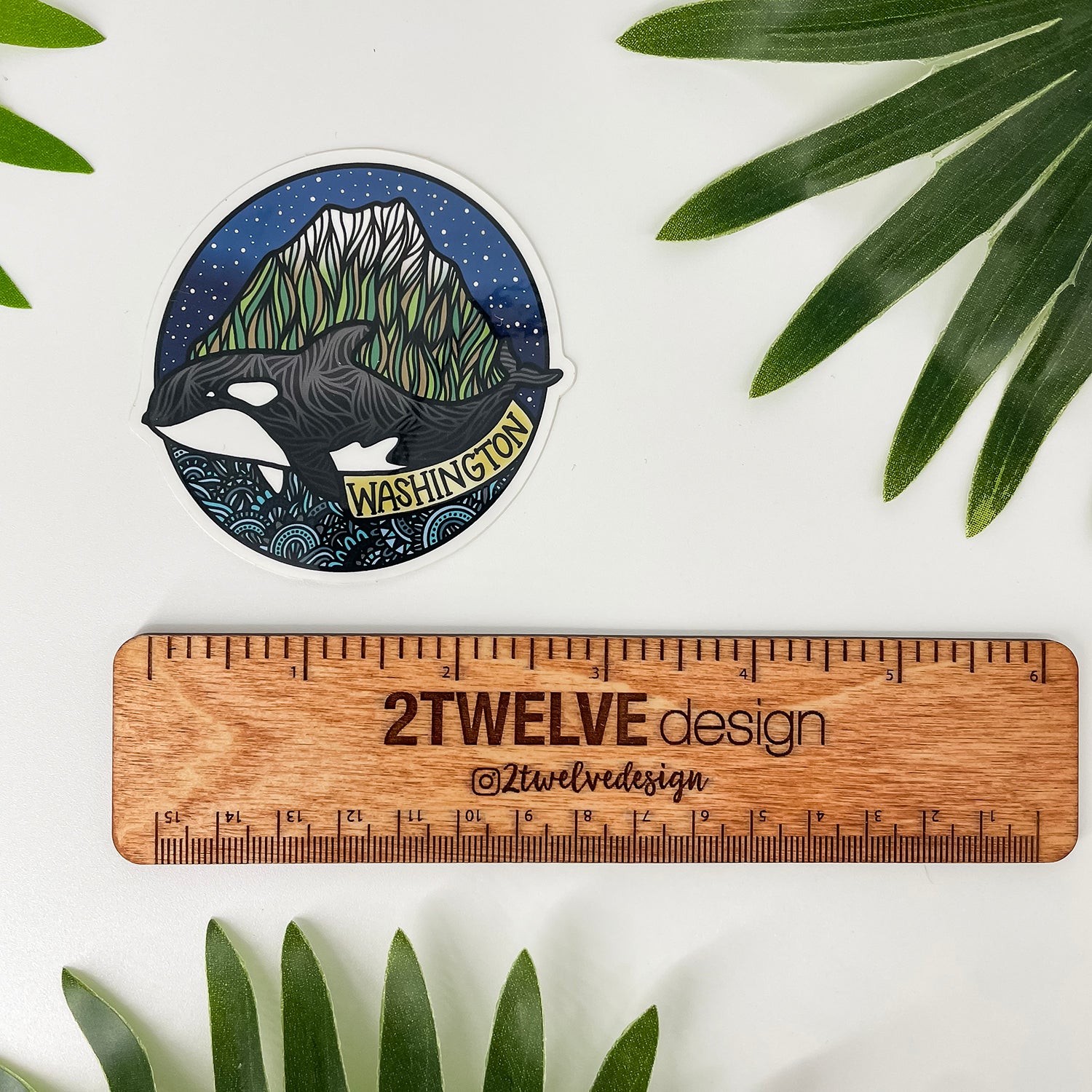 A circular sticker featuring an orca whale in front of a mountain range labeled 'WASHINGTON' is displayed on a white surface next to a wooden ruler and green palm leaves. The sticker is branded with '2Twelve Design'.
