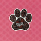 A hand-drawn paw print illustration in a black, red, and pink color palette, designed as a vinyl, waterproof die-cut sticker with the word 'Mom' incorporated into the design. Laying on a pink patterned background.