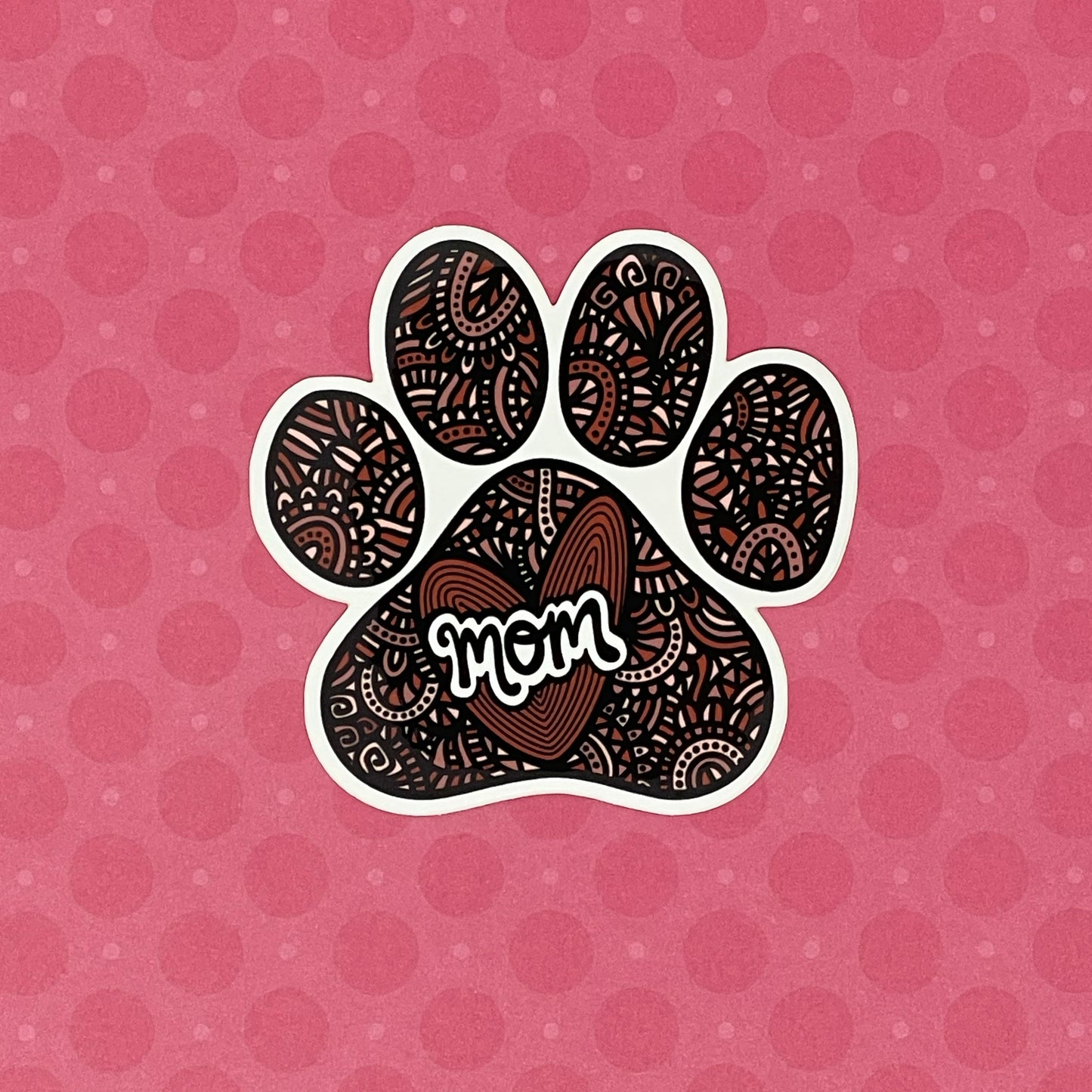 A hand-drawn paw print illustration in a black, red, and pink color palette, designed as a vinyl, waterproof die-cut sticker with the word 'Mom' incorporated into the design. Laying on a pink patterned background.