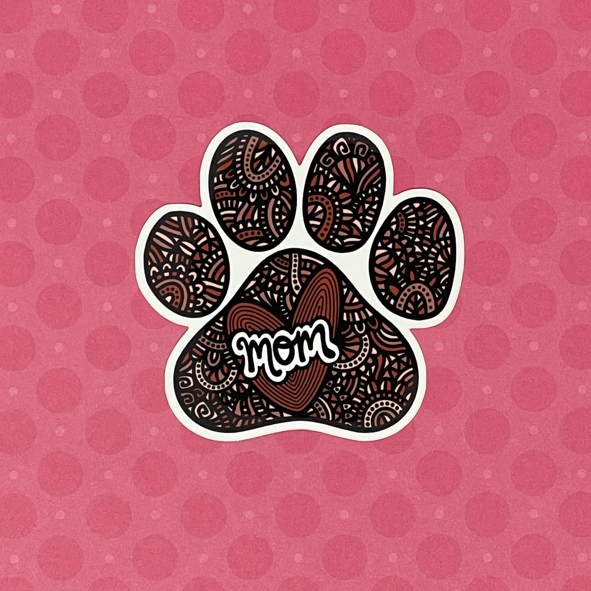 A hand-drawn paw print illustration in a black, red, and pink color palette, designed as a vinyl, waterproof die-cut sticker with the word 'Mom' incorporated into the design. Laying on a pink patterned background.