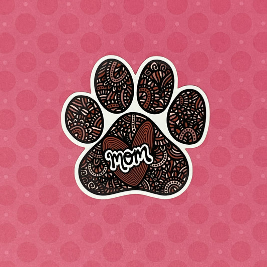 A hand-drawn paw print illustration in a black, red, and pink color palette, designed as a vinyl, waterproof die-cut sticker with the word 'Mom' incorporated into the design. Laying on a pink patterned background.