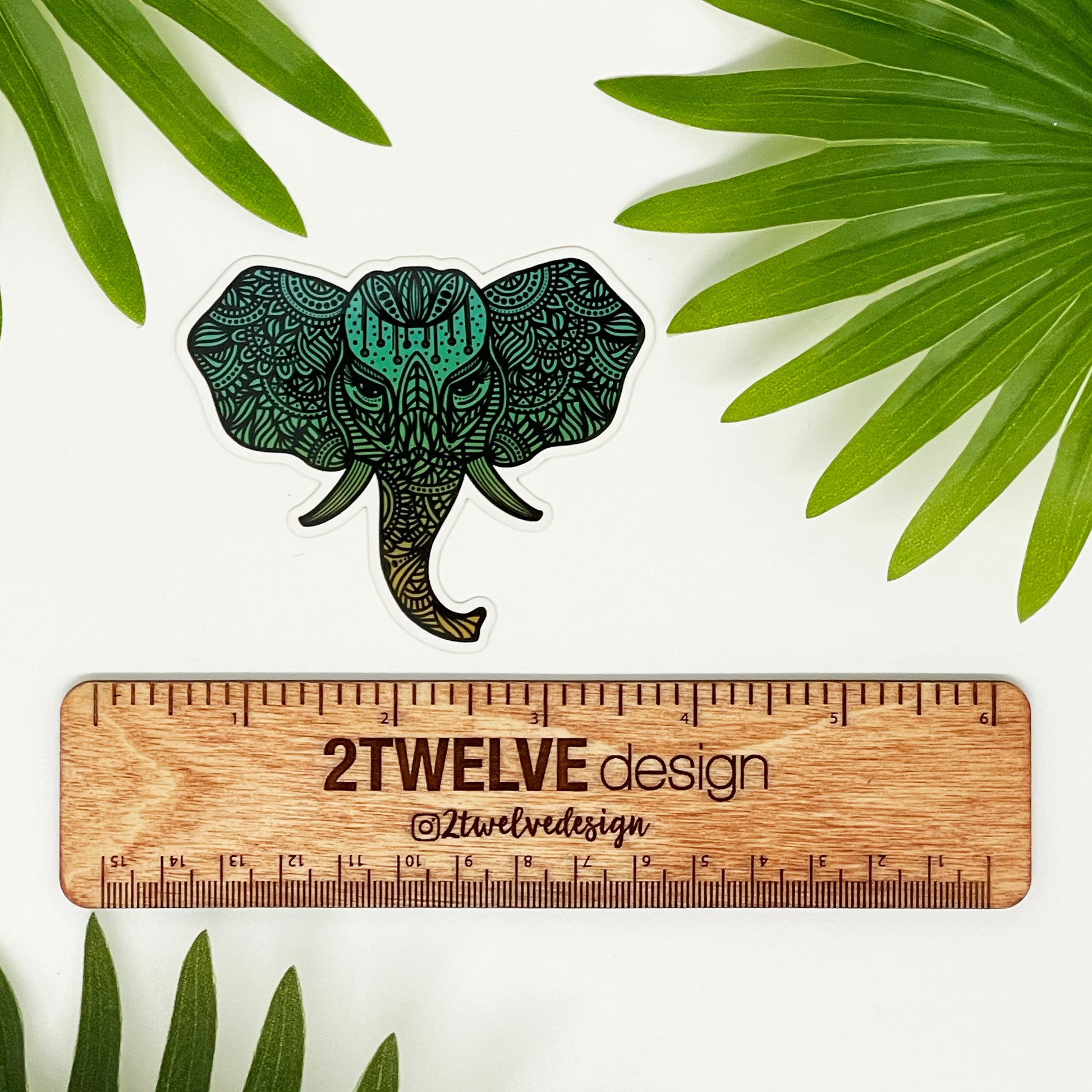 A hand illustrated elephant with intricate details made into a vinyl sticker. Laying on a white surface above a wooden "2Twelve Design" ruler. Surrounded by palm leaves