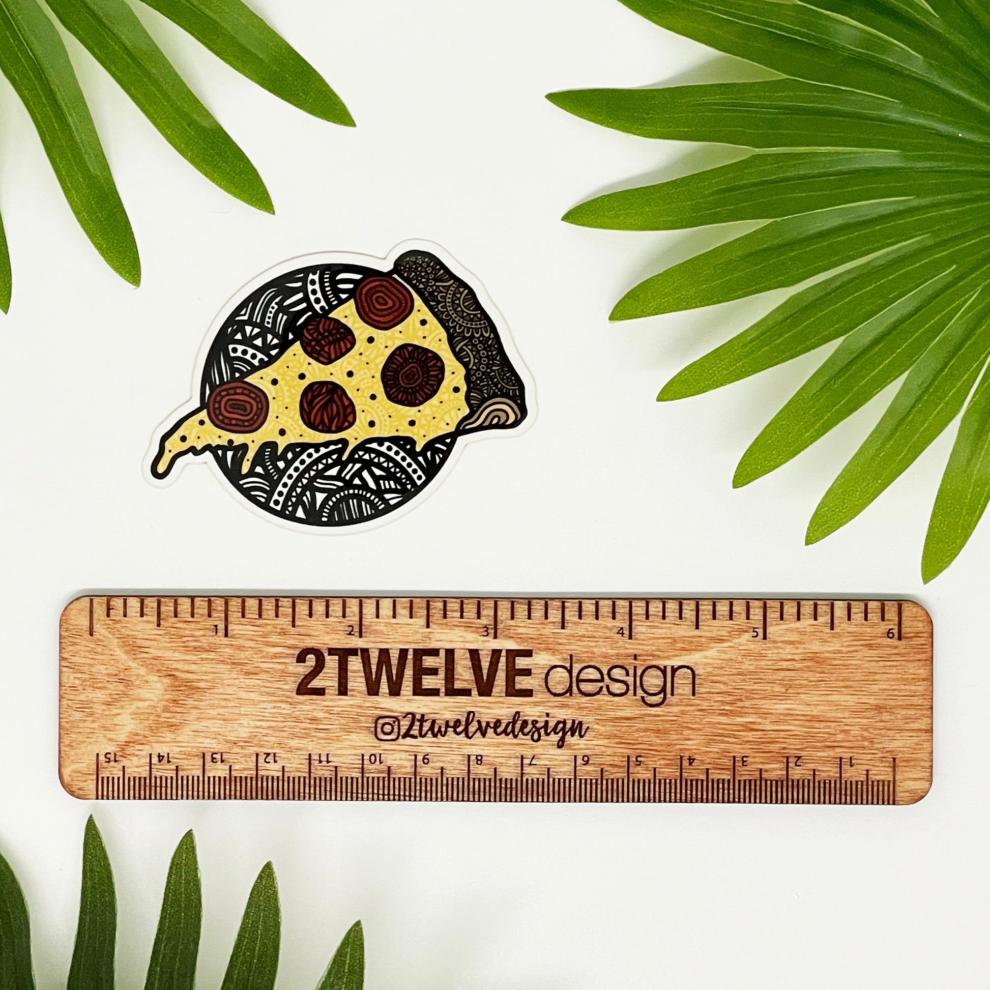 A hand illustrated pizza drawing made into a vinyl sticker. Laying on a white background surrounded by green palm leaves.