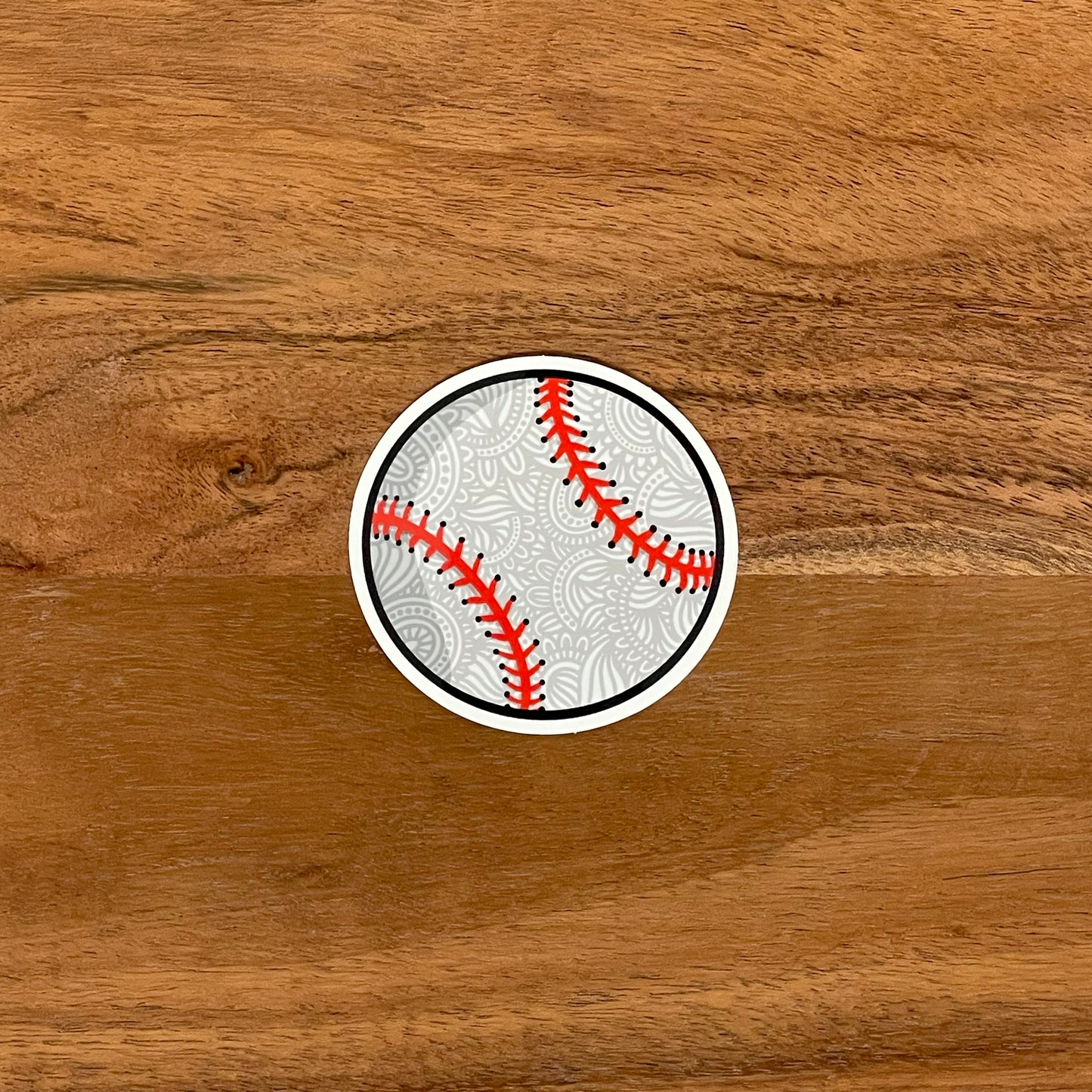 An illustrated baseball turned into a small sticker. Laying flat on a wooden surface.