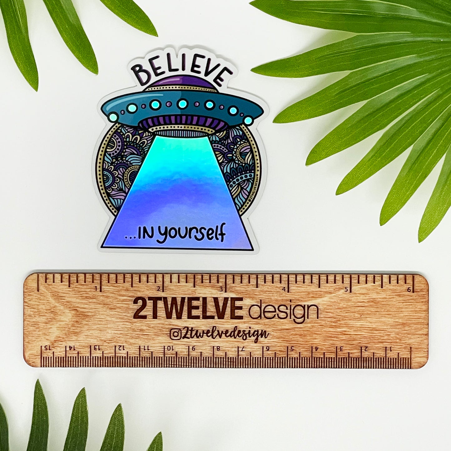 A holographic sticker of a spaceship with the quote "Believe In Yourself" laying on a white background surrounded by leaves with a ruler to show actual size.