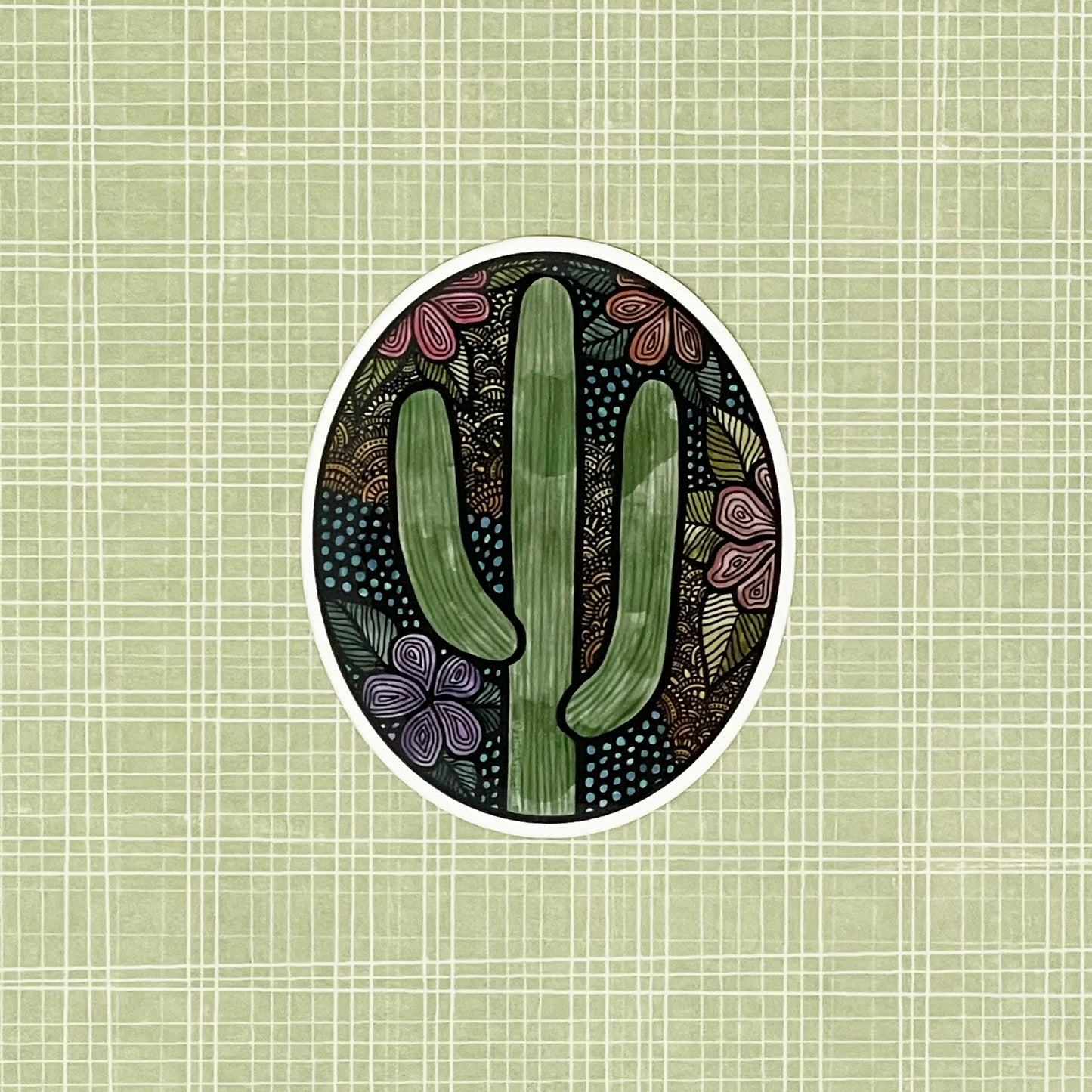 A cute floral, cactus illustration made into an oval vinyl sticker. Laying on a patterned green background.
