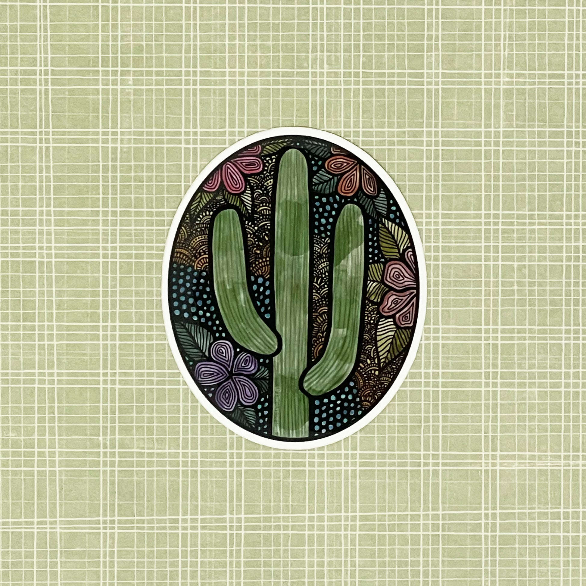 A cute floral, cactus illustration made into an oval vinyl sticker. Laying on a patterned green background.