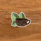 A die-cut vinyl sticker featuring a hand-drawn illustration of a coffee mug with monstera leaves on a wooden surface.