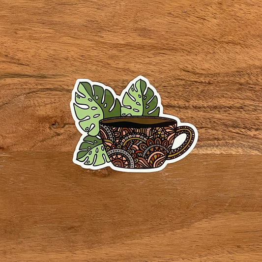 A die-cut vinyl sticker featuring a hand-drawn illustration of a coffee mug with monstera leaves on a wooden surface.
