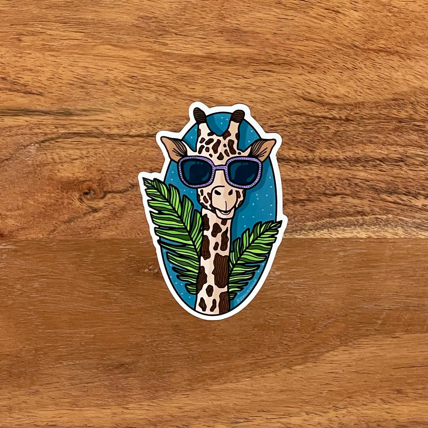 A stylin' giraffe illustration made into a vinyl sticker. Laying flat on a wooden surface.