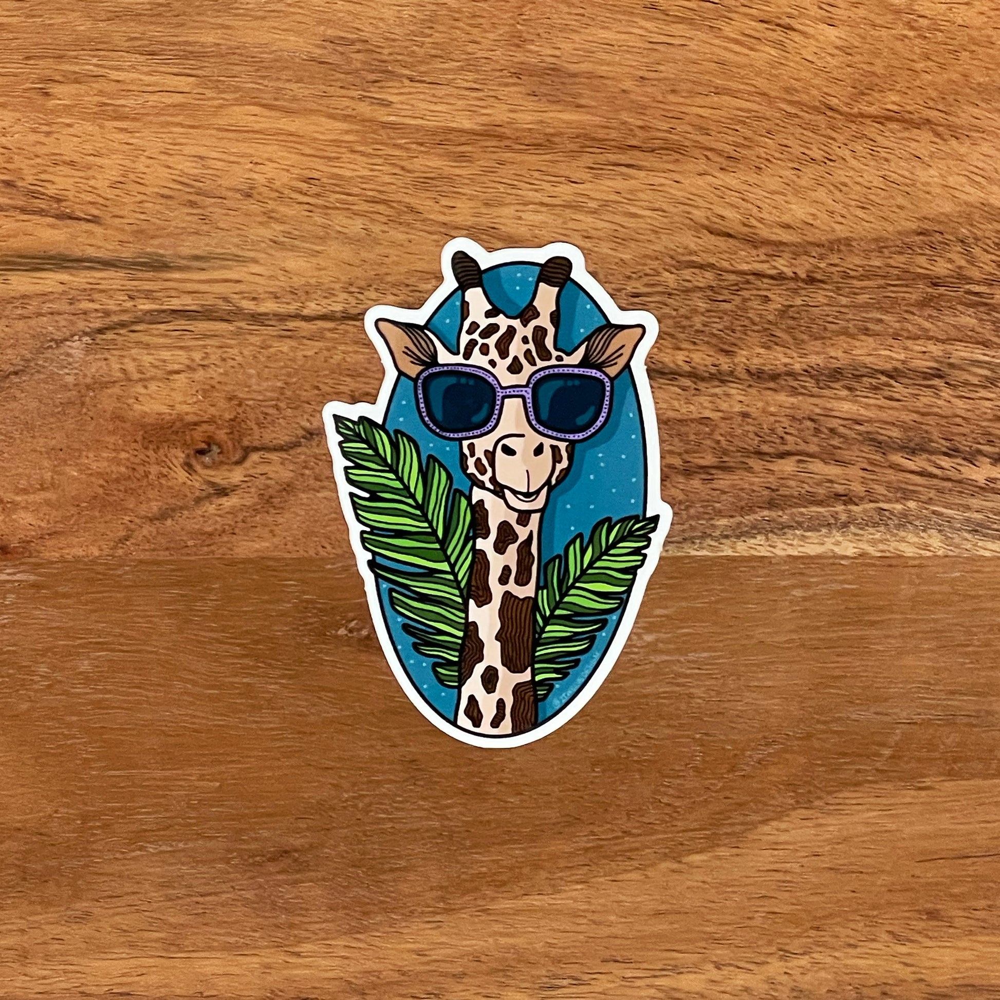 A stylin' giraffe illustration made into a vinyl sticker. Laying flat on a wooden surface.
