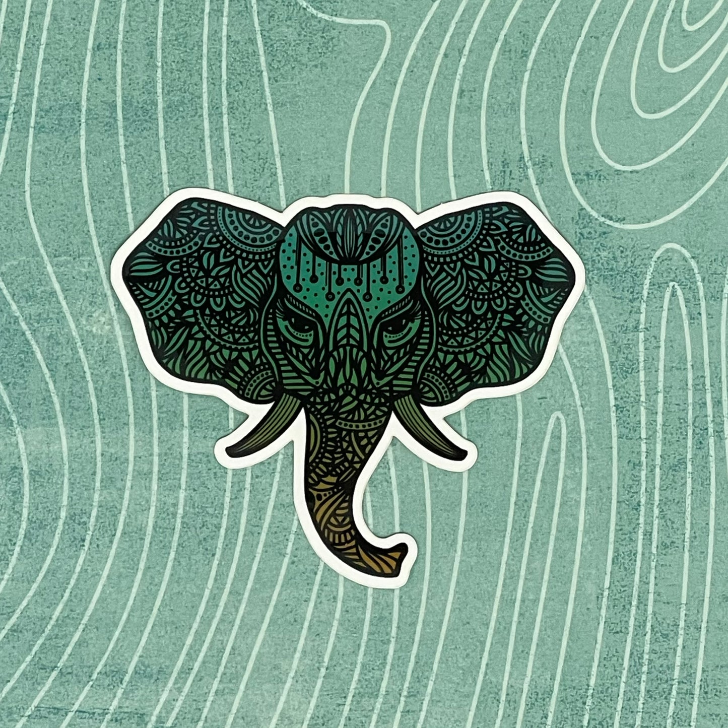 A teal colored, detailed elephant illustration made into a diecut sticker. Laying on a patterned background.