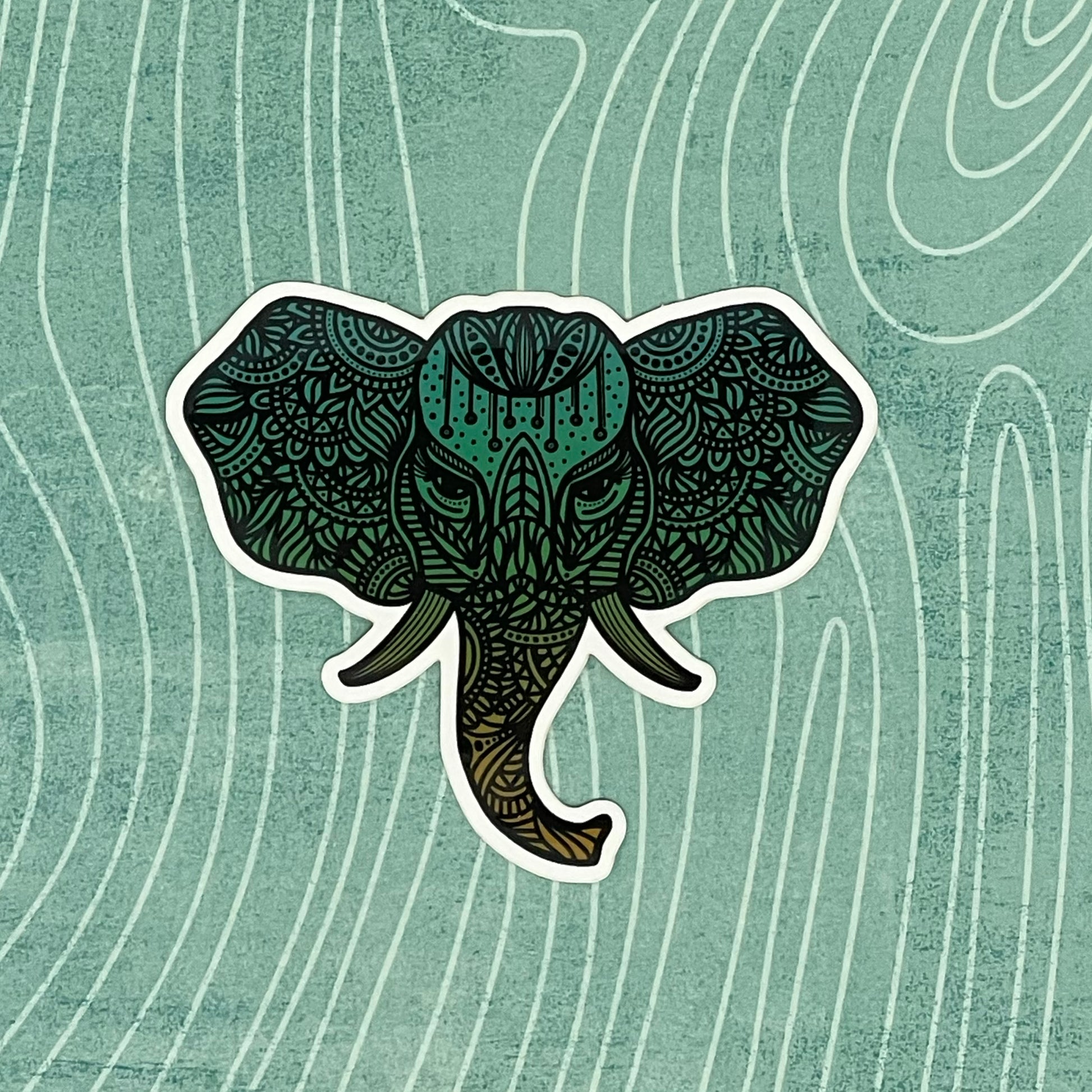 A teal colored, detailed elephant illustration made into a diecut sticker. Laying on a patterned background.