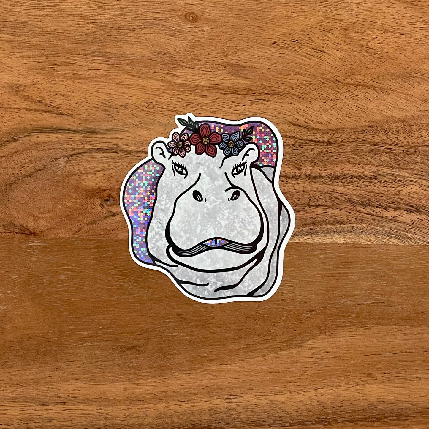 Cute hippo illustration made into a vinyl, glittery sticker. Laying on a wooden surface.