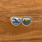 An illustration of sunglasses, sea and mountain design turned into a vinyl sticker. Laying on a wooden background.