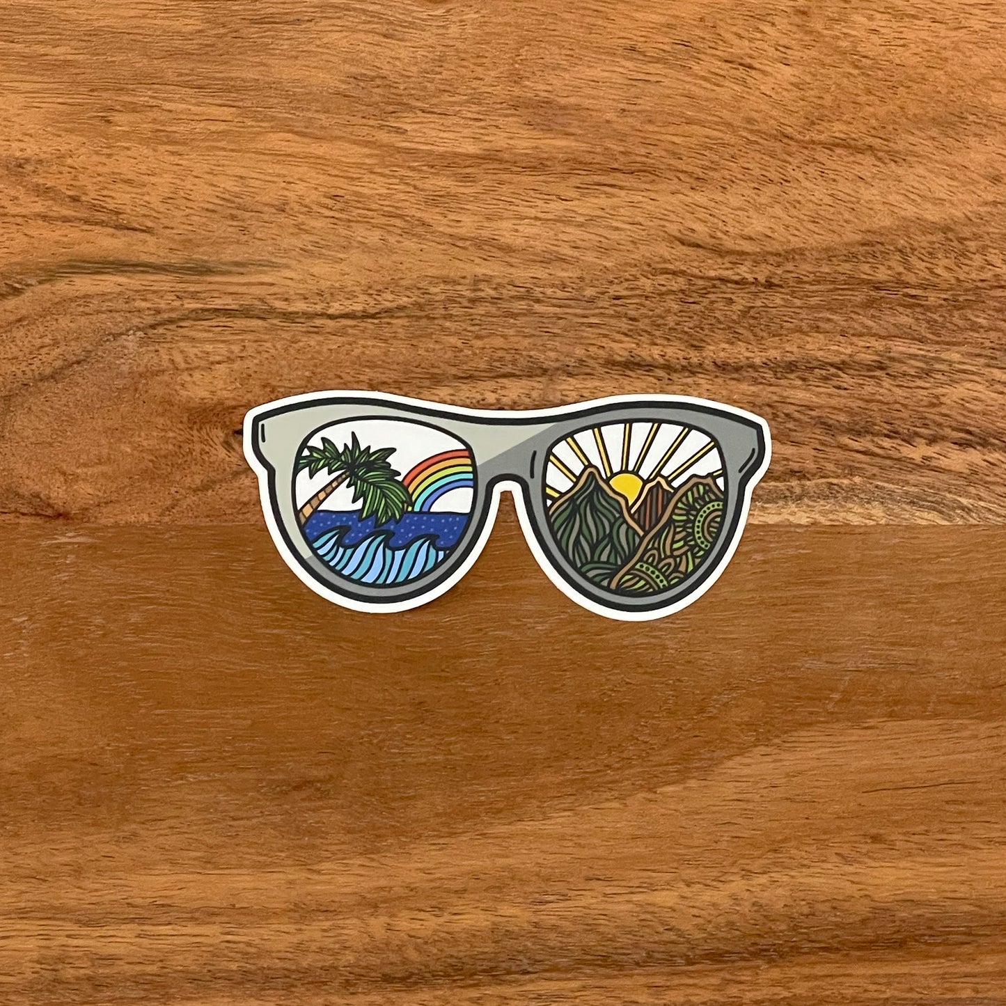 An illustration of sunglasses, sea and mountain design turned into a vinyl sticker. Laying on a wooden background.