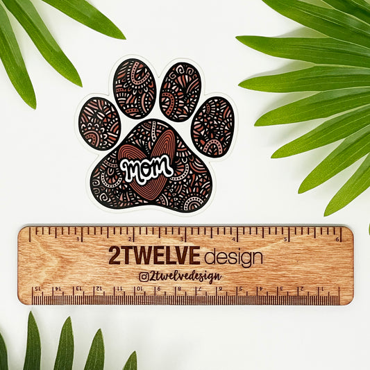 A hand-drawn paw print illustration in a black, red, and pink color palette, designed as a vinyl, waterproof die-cut sticker with the word 'Mom' incorporated into the design. Laying on a white background with a "2Twelve Design" ruler to show size. All surrounded by green palm leaves.