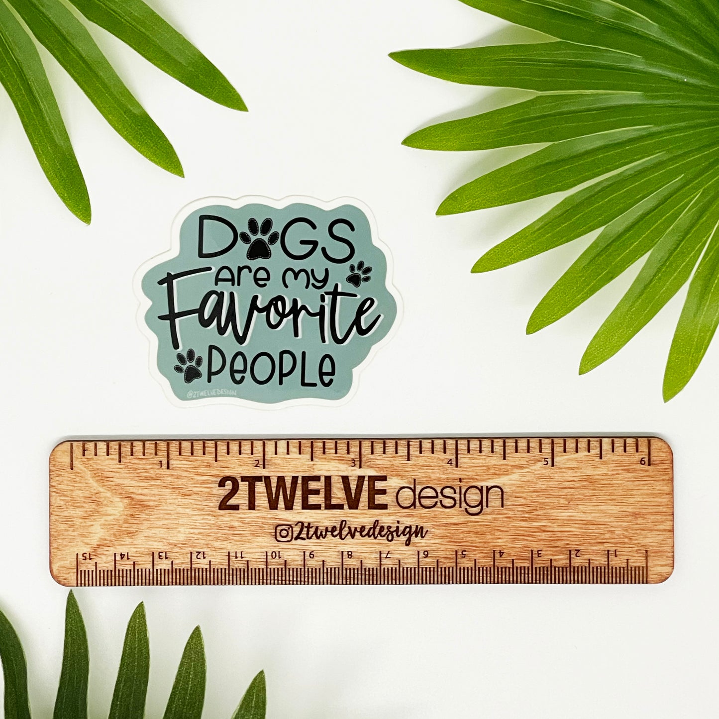 A sticker that says "Dogs are my Favorite People" with paw prints laying on a white surface surrounded by leaves and a ruler to show real size.