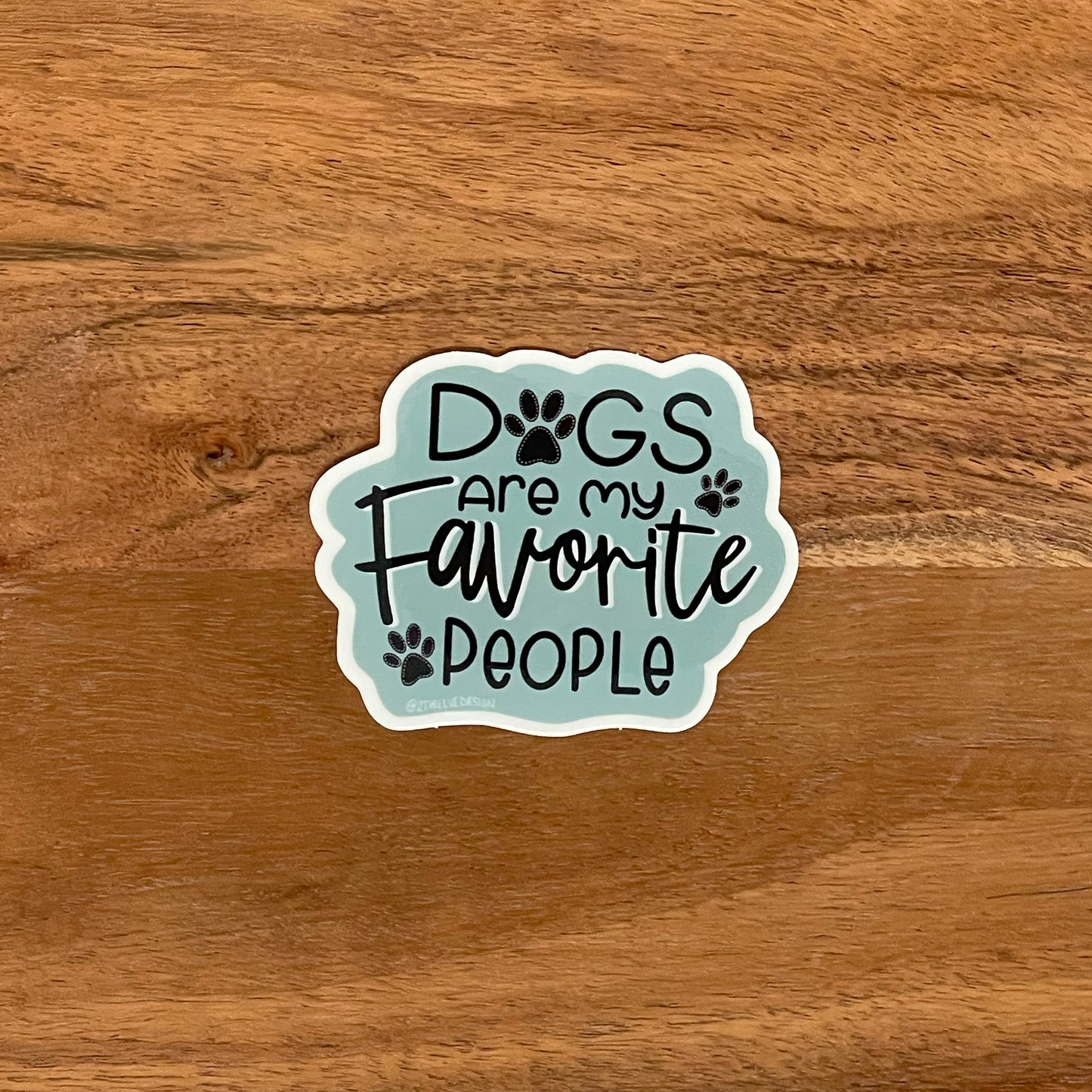 "Dogs are my favorite people" illustrated for a sticker with paw prints. Laying on a wooden surface.