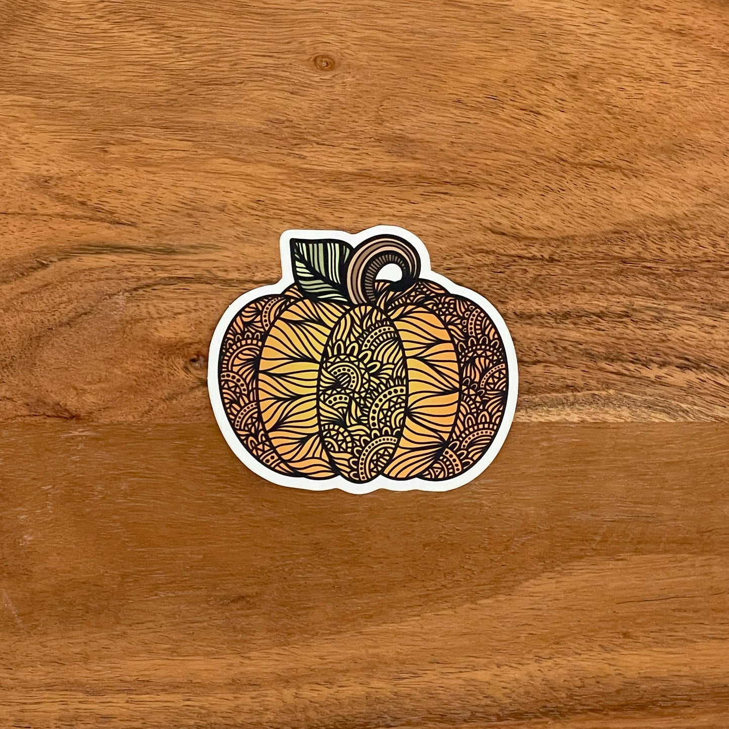 A cute, autumn pumpkin illustration with intricate details turned into a vinyl sticker. Laying on a wooden surface.