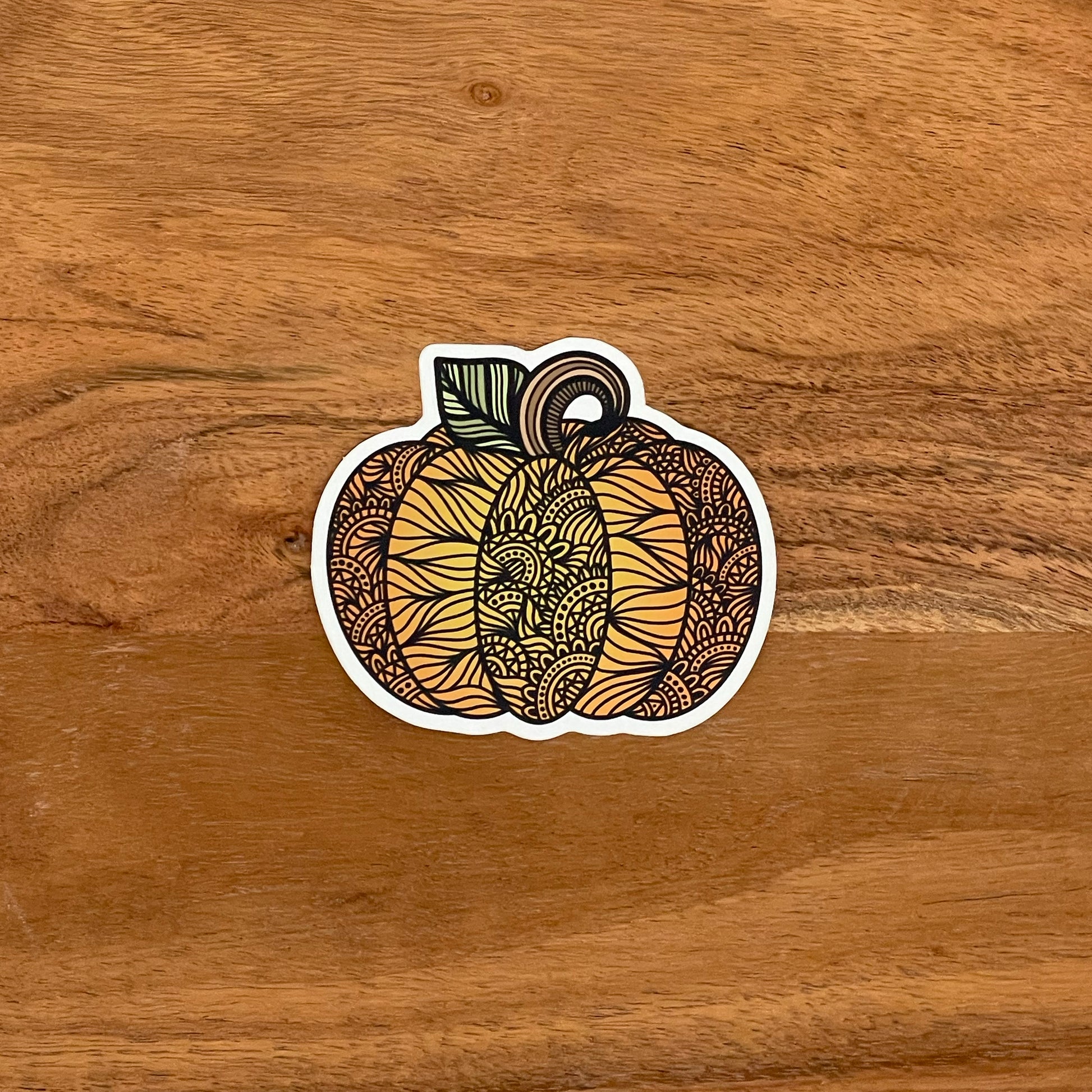A cute, autumn pumpkin illustration with intricate details turned into a vinyl sticker. Laying on a wooden surface.