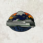 An illustration of a Washington State Ferry in the water with a mountain in the background - made into a vinyl sticker. Laying on a patterned gray surface.