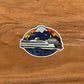 An illustrated ferry at sunset. Durable vinyl sticker laying on a wooden surface.