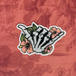 A vinyl sticker featuring a hand-drawn illustration of a skeleton shaka with floral elements and a matte shimmer finish, placed on a textured red background.