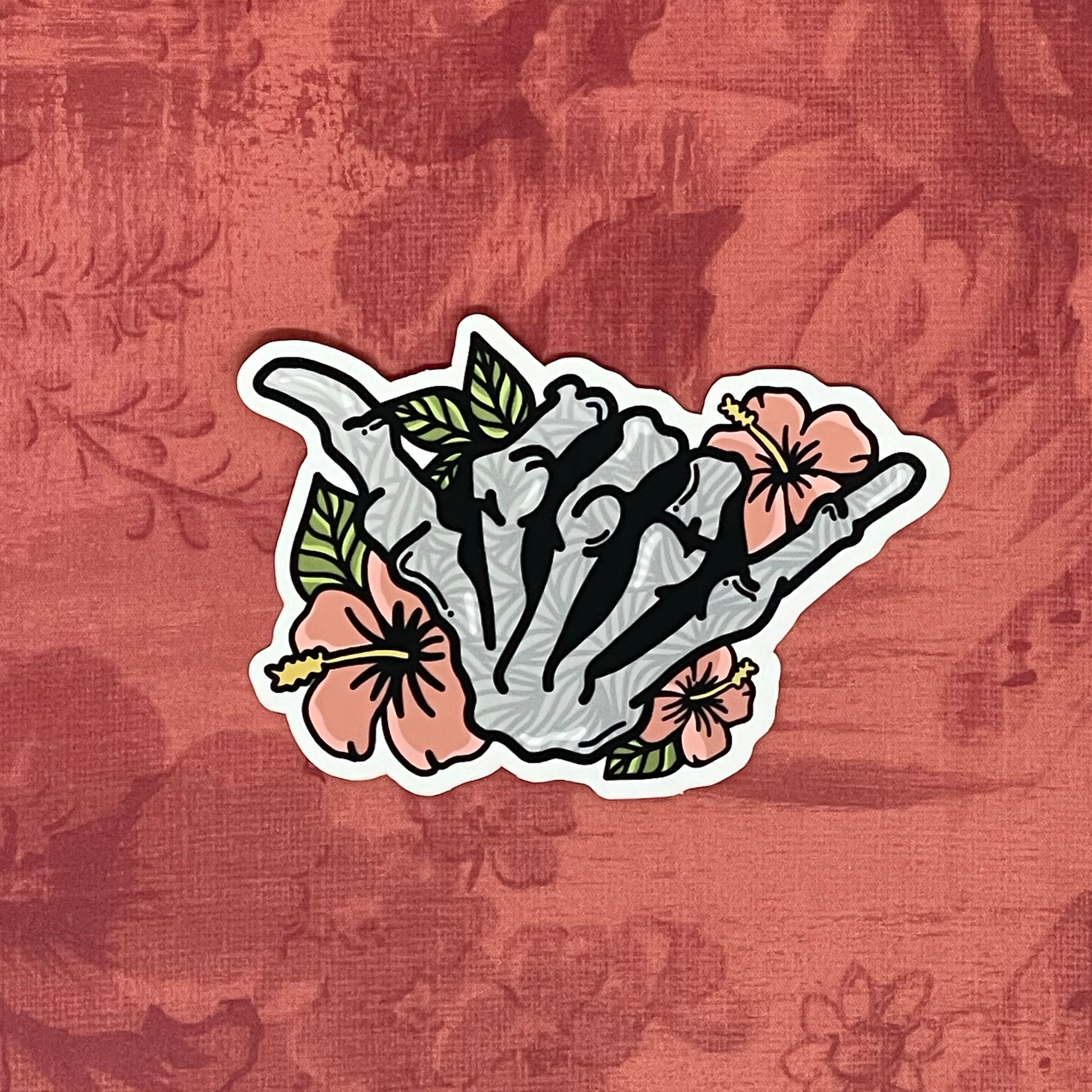 A vinyl sticker featuring a hand-drawn illustration of a skeleton shaka with floral elements and a matte shimmer finish, placed on a textured red background.