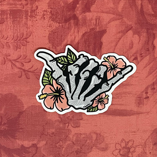 A vinyl sticker featuring a hand-drawn illustration of a skeleton shaka with floral elements and a matte shimmer finish, placed on a textured red background.